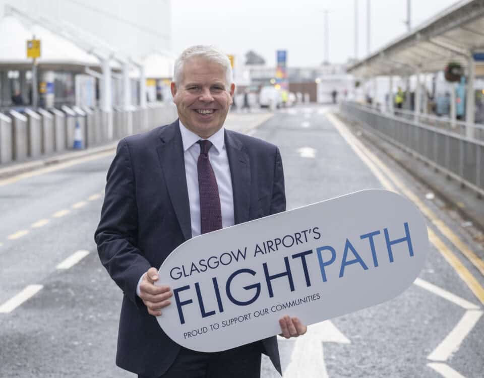 A man in a suit stands on a road outside an airport, smiling and holding a large sign that reads "Glasgow Airport's FLIGHTPATH – Proud to support our communities.