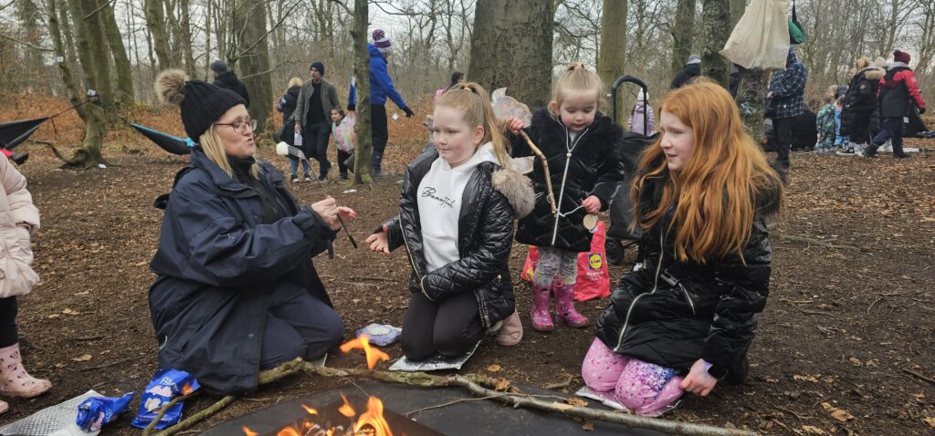 A group of children and an adult sit and kneel around a campfire in a forest, roasting marshmallows on sticks. Other people are seen in the background among the trees. The atmosphere is lively and social.