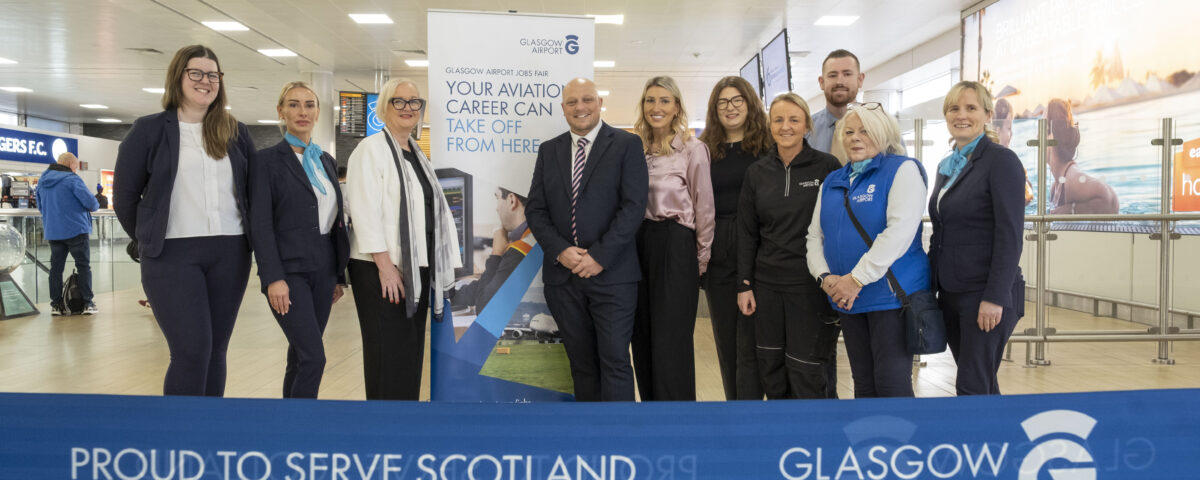 A group of ten people stand smiling behind a blue “Glasgow Airport: Proud to Serve Scotland” ribbon inside an airport terminal. A recruitment banner and check-in counters are visible in the background.