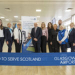 A group of ten people stand smiling behind a blue “Glasgow Airport: Proud to Serve Scotland” ribbon inside an airport terminal. A recruitment banner and check-in counters are visible in the background.
