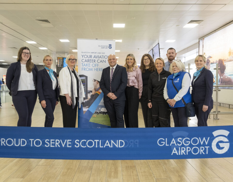 A group of ten people stand smiling behind a blue “Glasgow Airport: Proud to Serve Scotland” ribbon inside an airport terminal. A recruitment banner and check-in counters are visible in the background.