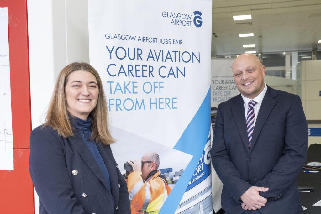 Two people in business attire stand smiling in front of a Glasgow Airport Jobs Fair banner that reads, "Your aviation career can take off from here," with an image of an airport worker and a control tower.