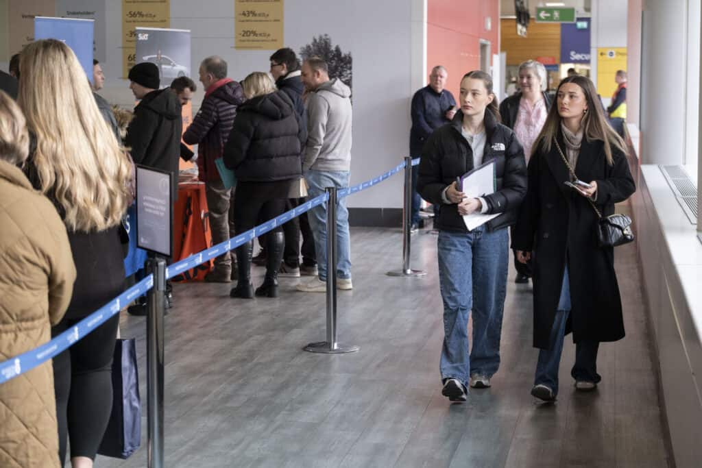 People stand in line at an indoor event. Two young women walk past the queue, one holding a notebook. Others are talking at tables with orange tablecloths. Signs and banners are visible in the background.