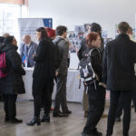 A group of people, some wearing backpacks and coats, stand and talk near display booths at an indoor event or career fair with posters and banners on the walls.
