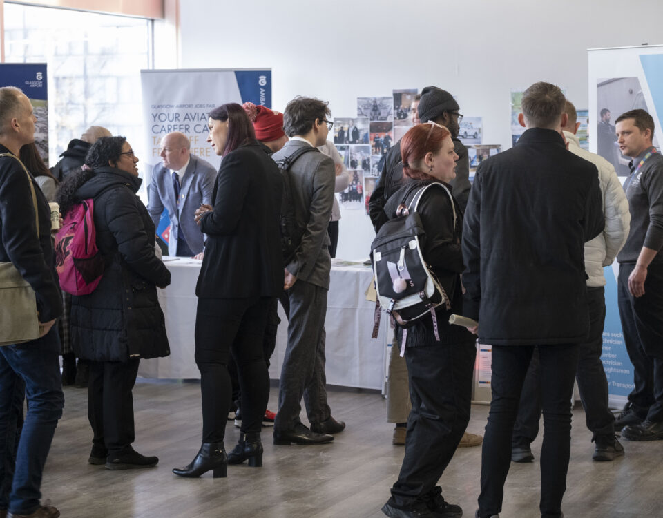 A group of people, some wearing backpacks and coats, stand and talk near display booths at an indoor event or career fair with posters and banners on the walls.