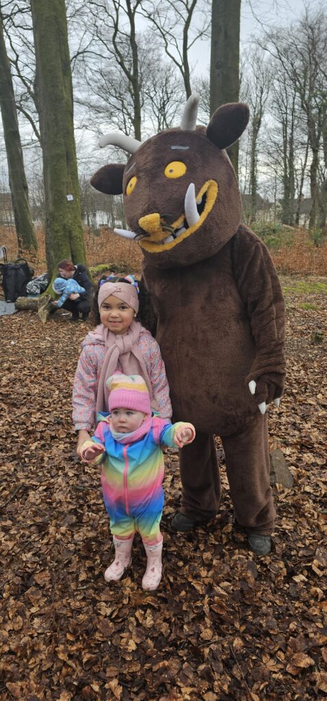 Two young children stand in a forest with a person in a Gruffalo costume. The older child smiles and hugs the younger, who wears a colorful outfit. Fallen leaves cover the ground and trees are in the background.