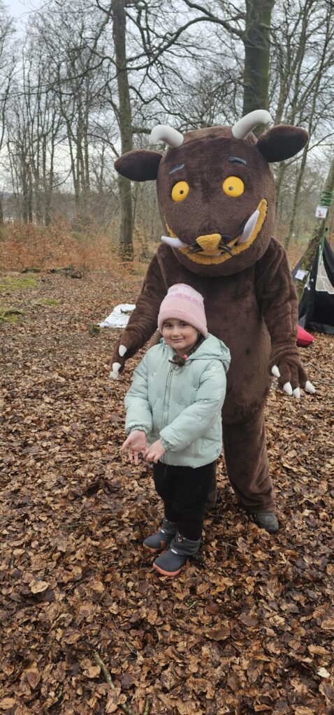 A young child in a pink hat and light blue coat stands smiling in front of a person wearing a large brown Gruffalo costume, outdoors on a leafy forest floor. Bare trees and overcast sky are in the background.