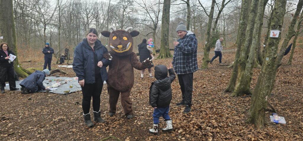 A person in a brown Gruffalo costume stands in a forest surrounded by people, including a woman smiling and a child reaching out. Other people are scattered in the background among bare trees and fallen leaves.