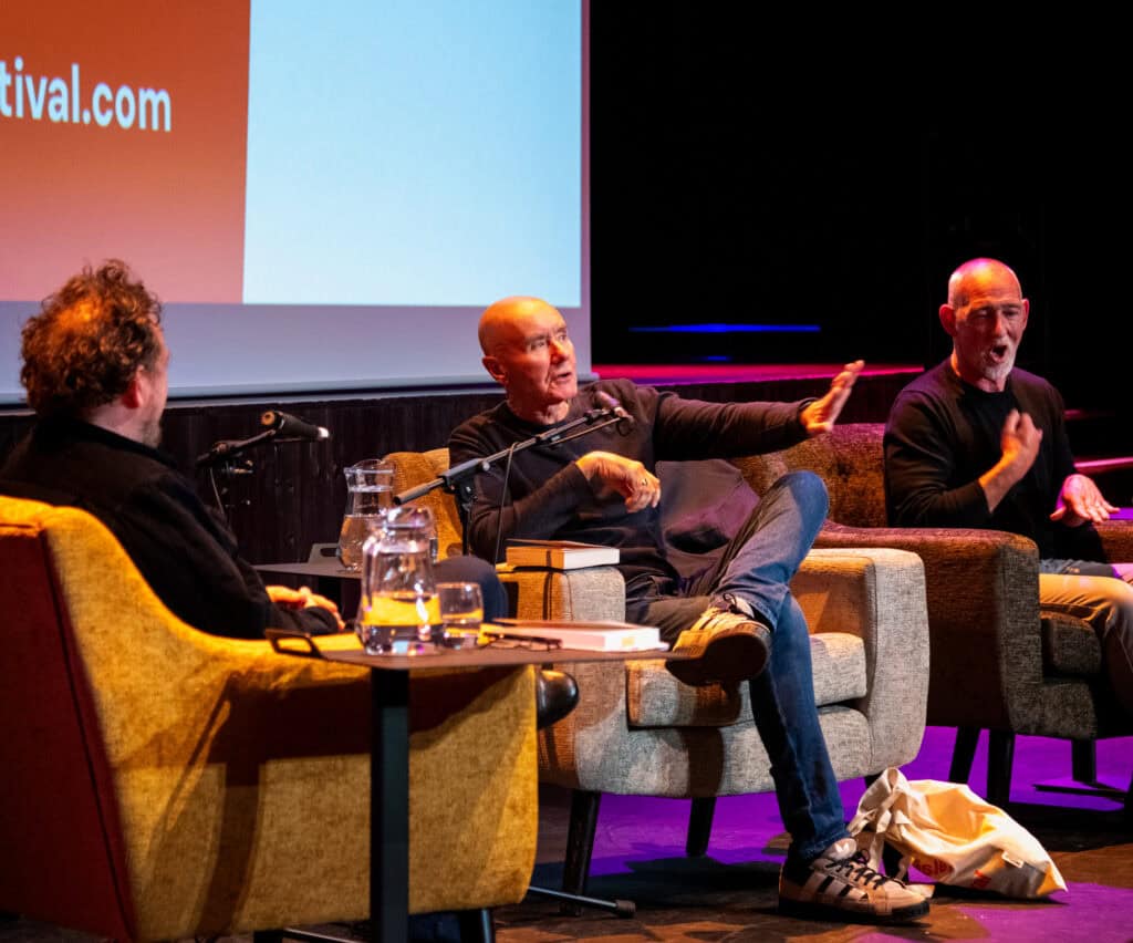 Three men sit on stage in armchairs, engaged in a lively discussion. One man gestures passionately with his hand, while the others listen. A table with water pitchers and microphones is in front of them. The background shows a large screen.