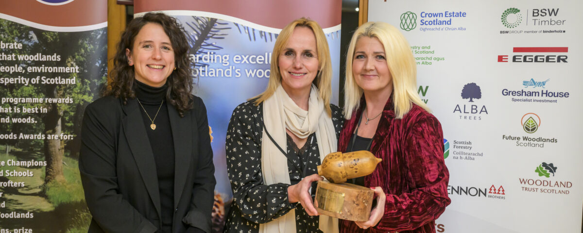 Three women stand together at an awards event. Two hold a wooden trophy. Banners behind them read “Scotland’s Finest Woods Awards” and list sponsors. All are smiling and dressed in business attire.