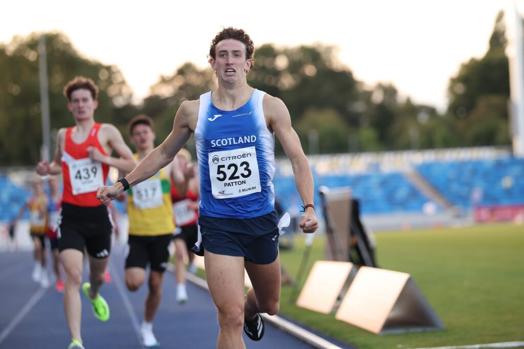 A male athlete in a blue Scotland uniform, number 523, celebrates as he crosses the finish line during a track race, with competitors trailing behind him on a stadium track.
