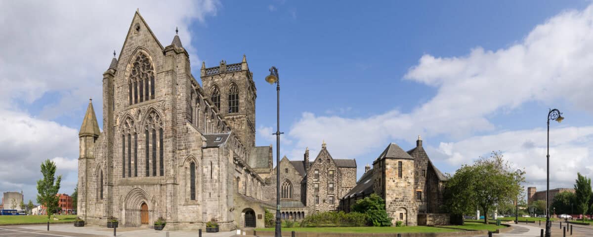 A wide-angle view of a large, historic stone cathedral with tall arched windows, surrounded by smaller stone buildings, trees, and a paved square under a partly cloudy blue sky.
