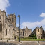 A wide-angle view of a large, historic stone cathedral with tall arched windows, surrounded by smaller stone buildings, trees, and a paved square under a partly cloudy blue sky.