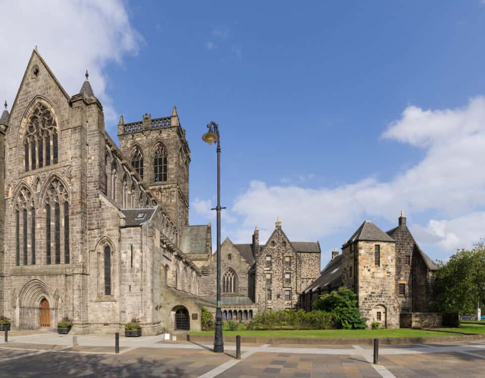 A wide-angle view of a large, historic stone cathedral with tall arched windows, surrounded by smaller stone buildings, trees, and a paved square under a partly cloudy blue sky.