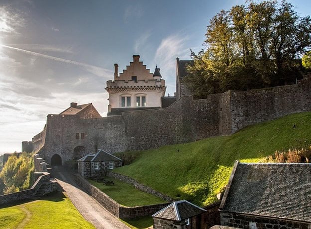 A historic stone castle with battlements and a white building stands atop a grassy hill, bordered by stone walls and surrounded by trees under a partly cloudy sky.