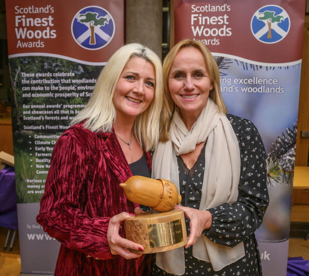 Two smiling women pose together holding a wooden acorn-shaped trophy at the Scotland's Finest Woods Awards. Banners in the background display the event’s name and logo, promoting excellence in Scottish woodlands.