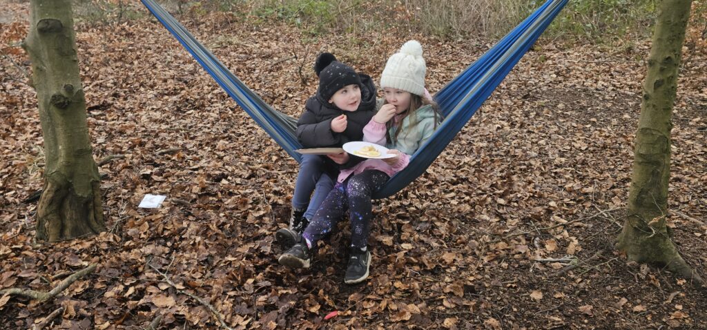Two children in winter hats and jackets sit together on a blue hammock between two trees, eating snacks from a plate. The ground is covered with brown fallen leaves in a woodland setting.