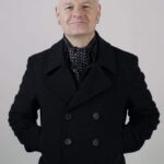 A man with short gray hair, wearing a black double-breasted coat and a black scarf with white dots, stands with his hands behind his back against a plain light background, smiling slightly at the camera.