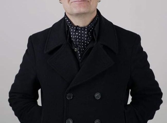 A man with short gray hair, wearing a black double-breasted coat and a black scarf with white dots, stands with his hands behind his back against a plain light background, smiling slightly at the camera.