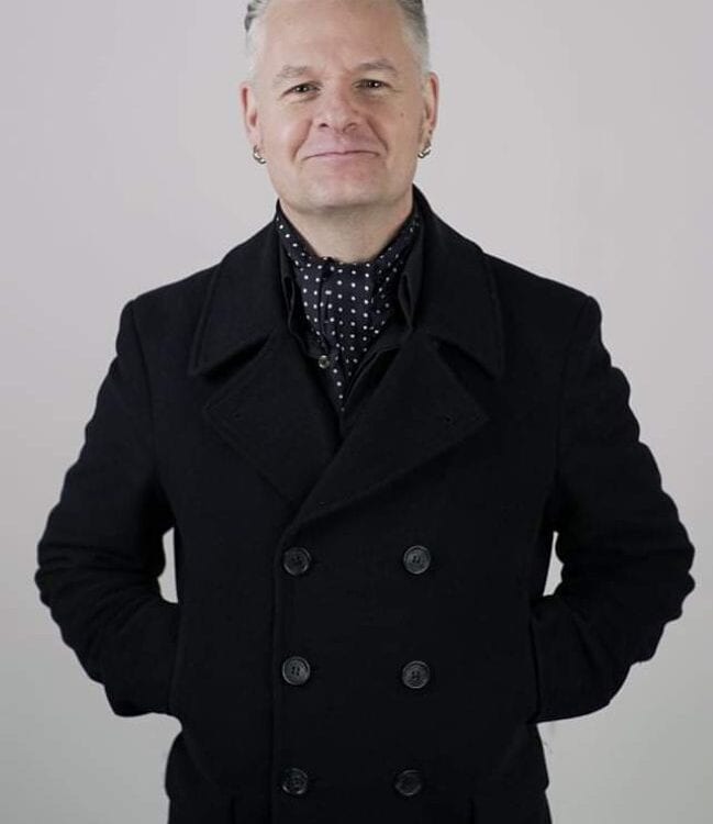 A man with short gray hair, wearing a black double-breasted coat and a black scarf with white dots, stands with his hands behind his back against a plain light background, smiling slightly at the camera.