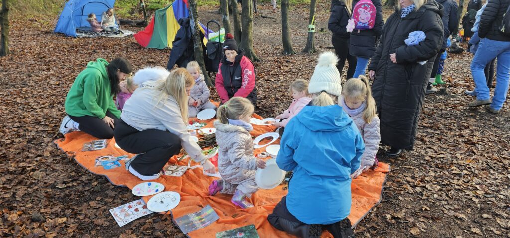 A group of children and adults sit and stand around an orange tarp outdoors, engaged in arts and crafts activities. Painted plates, leaves, and art supplies are scattered on the ground. Trees and fallen leaves surround the scene.
