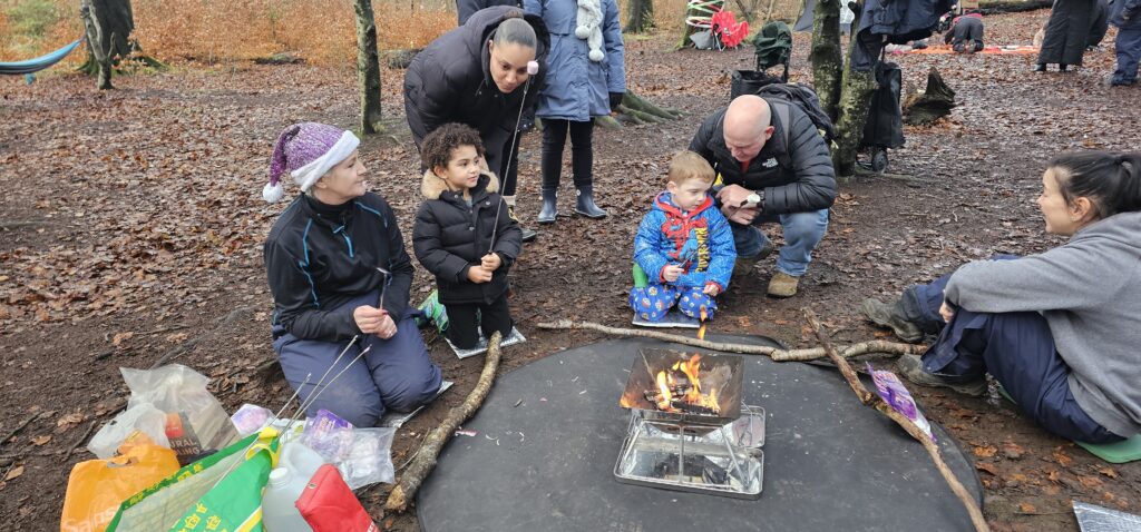A group of adults and young children sit around a small campfire in a wooded area, bundled up in warm clothing. Leaves cover the ground and supplies are scattered nearby. Everyone looks engaged and happy.