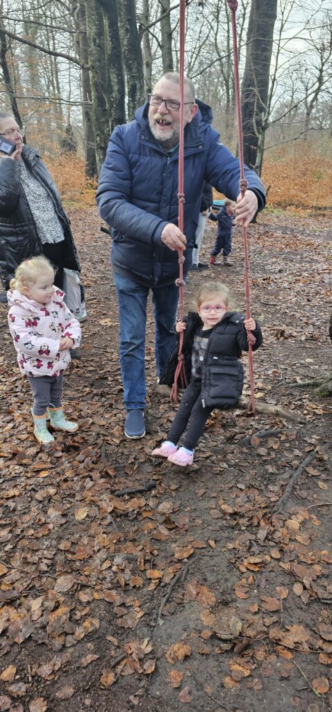 A man pushes a young girl on a swing in a forested park while another child stands nearby. Fallen leaves cover the ground, and several people are visible in the background. It is a cloudy day.