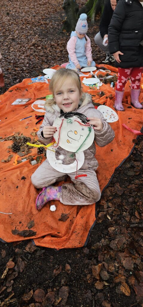 A young child in a beige snowsuit sits on an orange blanket outdoors, smiling and holding up a handmade paper plate decoration. Other children and art supplies are scattered around on the blanket, with autumn leaves on the ground.