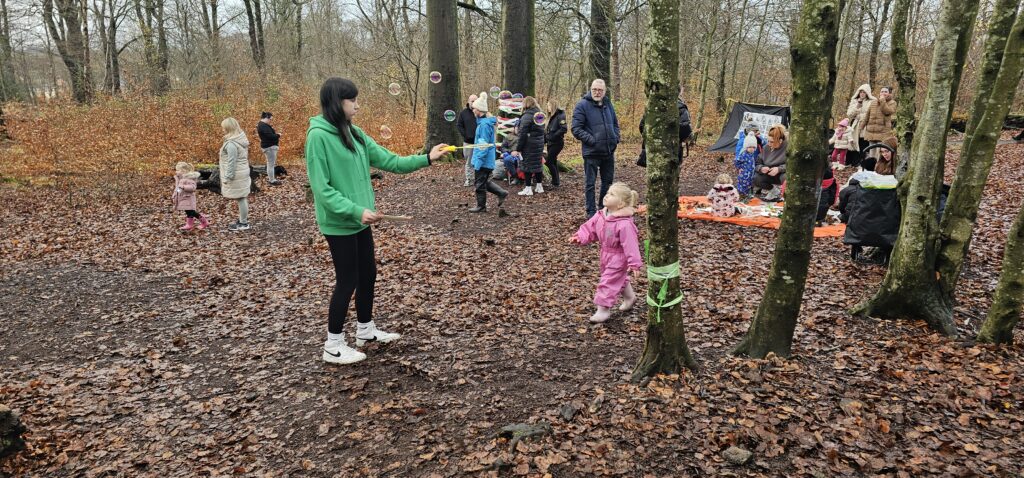 A woman in a green jacket blows bubbles for a young child in pink overalls in a wooded area. Several adults and children watch or play in the background among autumn leaves.