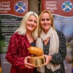 Two women smiling and holding a wooden acorn-shaped award, standing in front of banners for Scotland’s Finest Woods Awards.