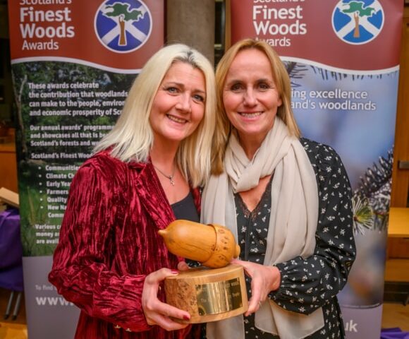 Two women smiling and holding a wooden acorn-shaped award, standing in front of banners for Scotland’s Finest Woods Awards.