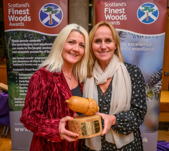 Two women smiling and holding a wooden acorn-shaped award, standing in front of banners for Scotland’s Finest Woods Awards.