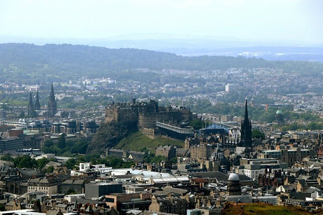 A panoramic view of Edinburgh, featuring Edinburgh Castle on a rocky hill surrounded by historic buildings, church spires, and a sprawling cityscape with green hills in the distance.