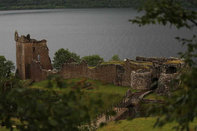 A stone castle ruin sits beside a lake, surrounded by green grass and trees. The remnants of a tall tower and winding walls are visible, with water and forested hills in the background.