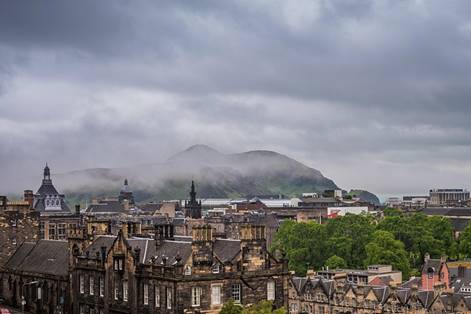 A cloudy, overcast sky looms above historic stone buildings in a city, with green hills and mist in the background.