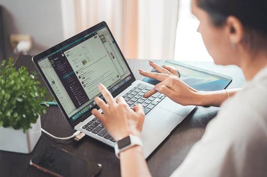 A woman types on a laptop displaying a messaging application, with a smartphone, tablet, and potted plant on the desk beside her. She wears a smartwatch and is working in a well-lit room.