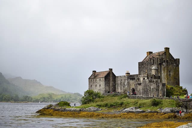 A historic stone castle stands on a grassy shore beside a body of water, with misty hills and cloudy skies in the background. Small groups of people are visible near the castle.