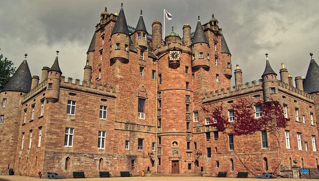 A large, historic stone castle with round towers, conical roofs, and crenellated walls stands under a cloudy sky. A flag flies from the central tower, and a clock is visible above the main entrance.