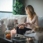 A woman with wavy blonde hair sits on a gray couch, wearing a cozy light sweater and black jeans. She faces a fluffy white dog, possibly a Samoyed, with candles and decor on a glass coffee table nearby.
