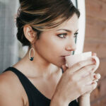 A woman with light brown hair in an updo is wearing a sleeveless black top and large earrings, holding a white mug close to her face as she sips, while gazing thoughtfully to the side.
