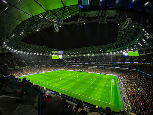 A brightly lit soccer stadium at night, packed with fans, shows a match in progress on a green pitch surrounded by stands under a large, curved roof.