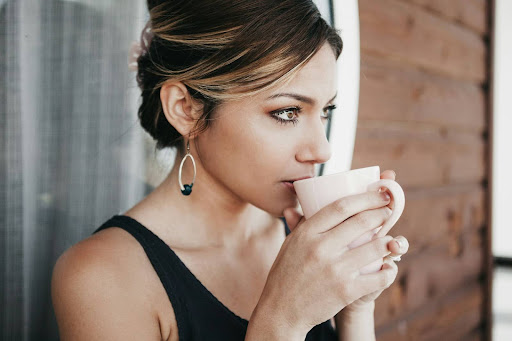 A woman with light brown hair in an updo is wearing a sleeveless black top and large earrings, holding a white mug close to her face as she sips, while gazing thoughtfully to the side.