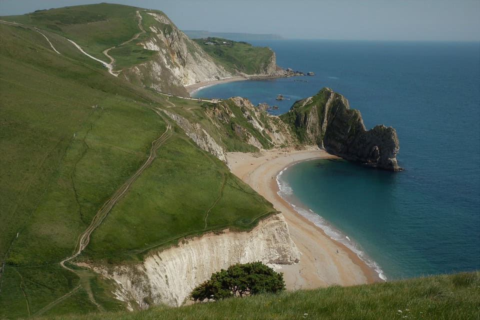 A coastal landscape with steep green cliffs, a curved sandy beach, and dramatic rock formations extending into calm blue sea under a clear sky.