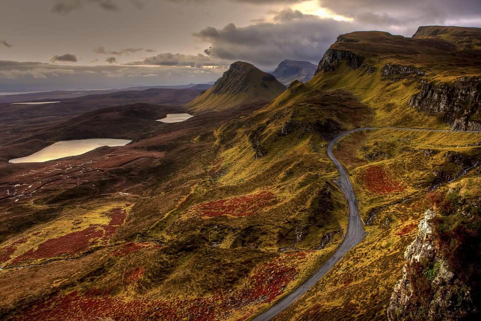 A winding road curves through rolling hills and rugged cliffs covered in green and red vegetation, with lakes and dramatic clouds in the background under a muted sky.