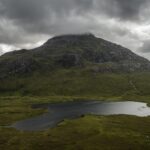 A dark, cloudy sky hangs over a green mountain partially covered by mist, with a small, reflective lake at its base surrounded by grassy terrain.