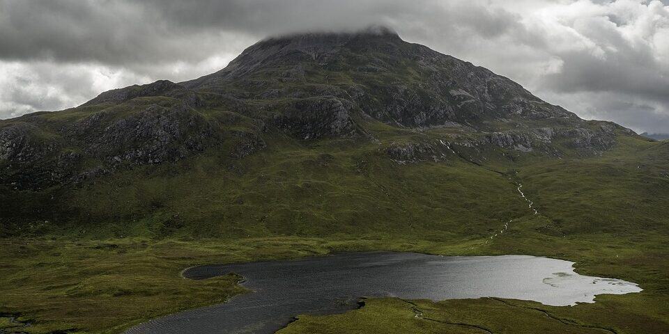 A dark, cloudy sky hangs over a green mountain partially covered by mist, with a small, reflective lake at its base surrounded by grassy terrain.