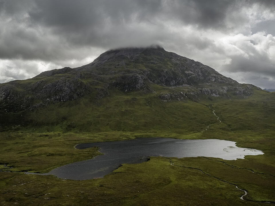A dark, cloudy sky hangs over a green mountain partially covered by mist, with a small, reflective lake at its base surrounded by grassy terrain.
