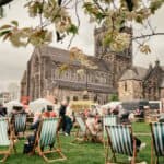 People relax on striped deck chairs at an outdoor event near a large historic stone church. Tents and stalls are set up on the grass, and flowering tree branches frame the scene. The sky is overcast.