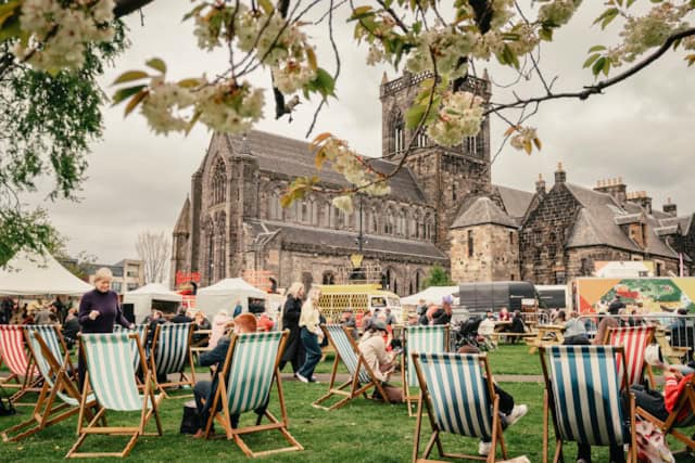 People relax on striped deck chairs at an outdoor event near a large historic stone church. Tents and stalls are set up on the grass, and flowering tree branches frame the scene. The sky is overcast.