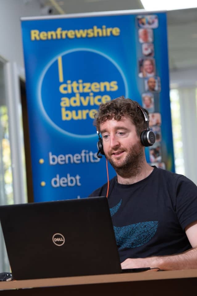 A man with curly hair wearing headphones sits at a desk using a laptop. Behind him is a Renfrewshire Citizens Advice Bureau banner listing “benefits” and “debt” among its services.